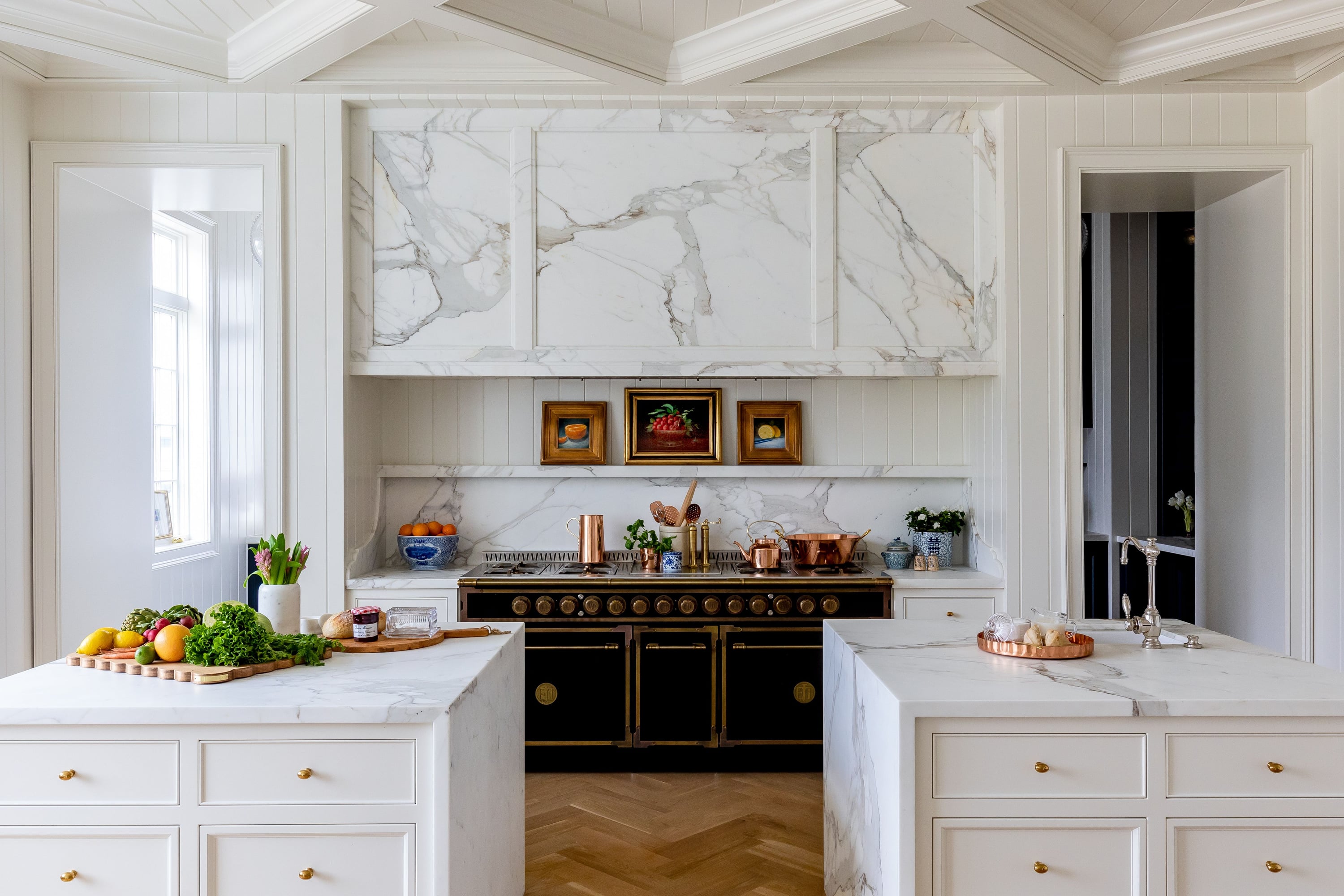 High-end white kitchen featuring dual marble islands, a black professional stove with brass knobs, and a massive marble range hood.